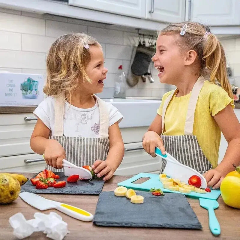 Two young girls in aprons laugh together while cutting fruit with a Soulful Trading Mini Chef Kid Friendly Cooking Toolset in a kitchen.