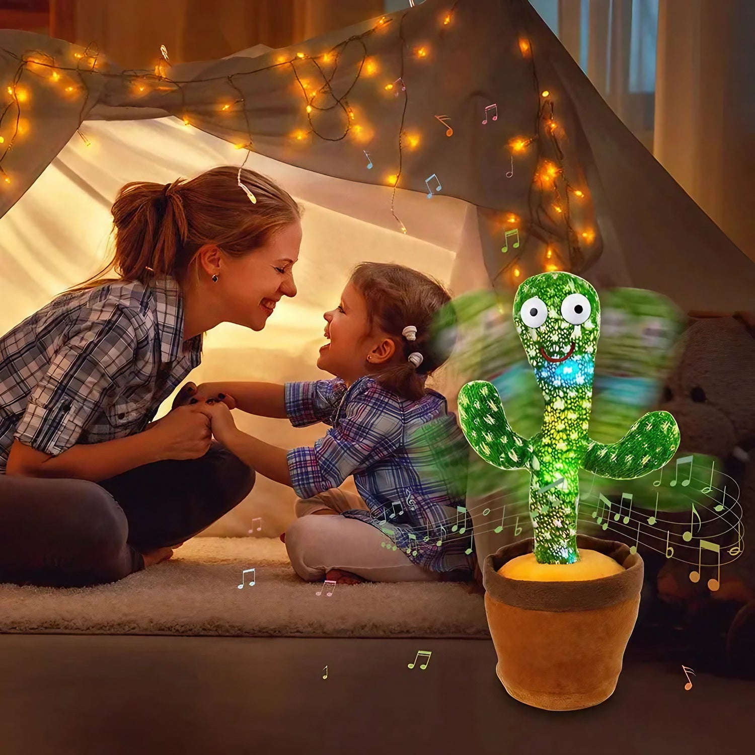 A woman and a young girl smiling at each other inside a cozy blanket fort lit by string lights, with an Original Dancing Cactus Toy from Soulful Trading nearby.