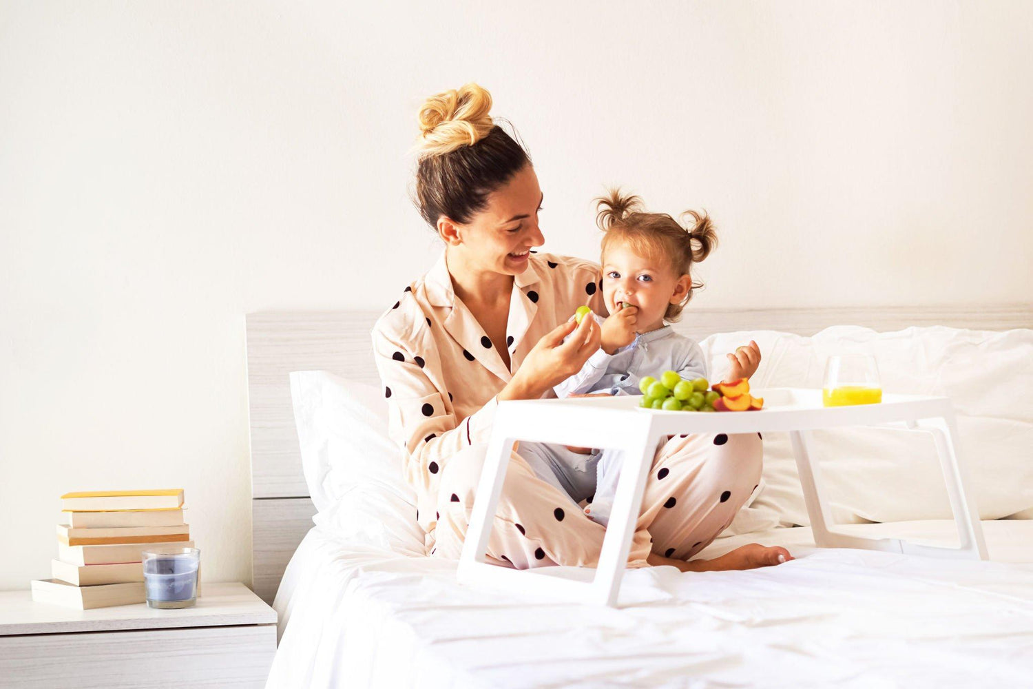 A mother in polka-dot pajamas feeding her toddler at a small white table on a bed, both smiling, with books and a juice glass nearby.
