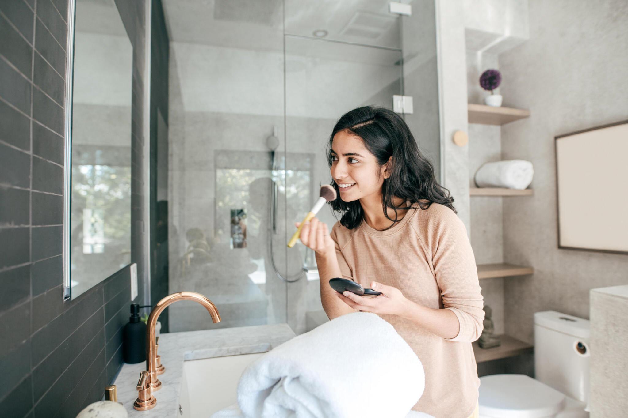 A woman brushes her teeth while looking at her reflection in a bathroom mirror, holding a smartphone in her other hand.