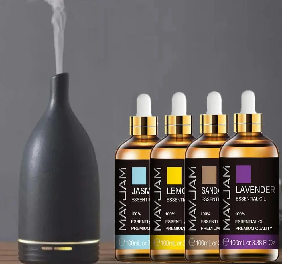 A black ultrasonic essential oil diffuser with visible mist next to four bottles of Soulful Trading Aromatic Essential Oils labeled may chang, lemongrass, sandalwood, and lavender.