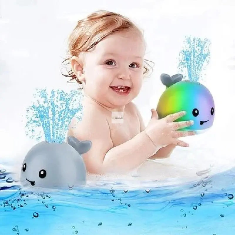 A cheerful baby playing with a colorful Soulful Trading Sprinkler Whale Bath Toy in water, splashes visible around.