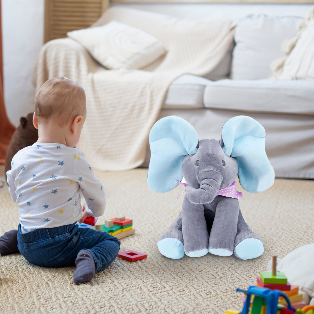 A baby sits on the floor facing a Peek a Boo Musical Elephant toy from Soulful Trading, surrounded by colorful blocks, in a cozy living room setting.