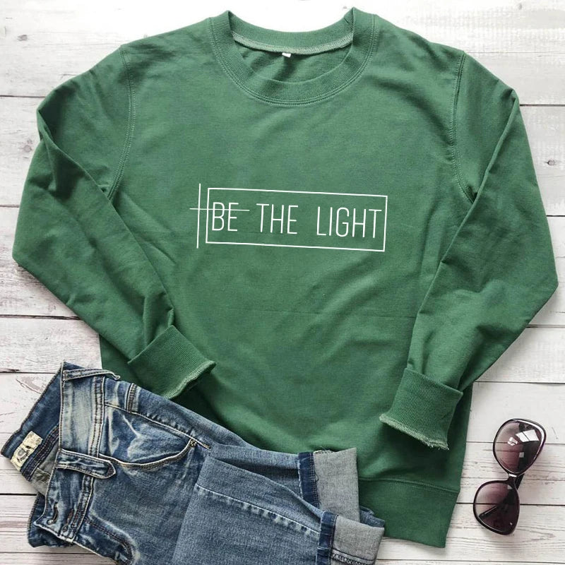 Green cotton Be The Light Christian sweatshirt from Soulful Trading, paired with jeans and sunglasses, displayed on a wooden surface.