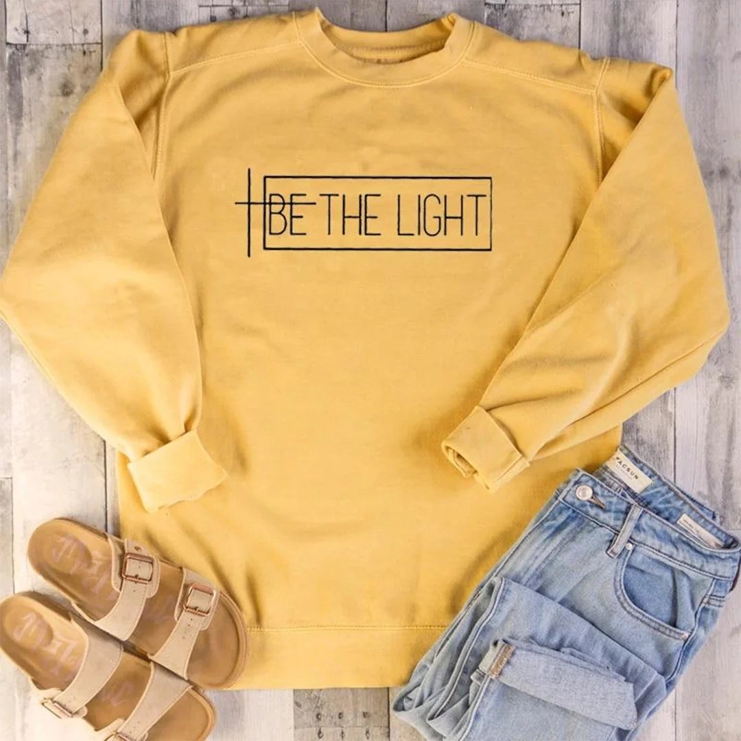 Yellow cotton Be The Light Christian sweatshirt from Soulful Trading, paired with blue jeans and brown sandals on a wooden floor.
