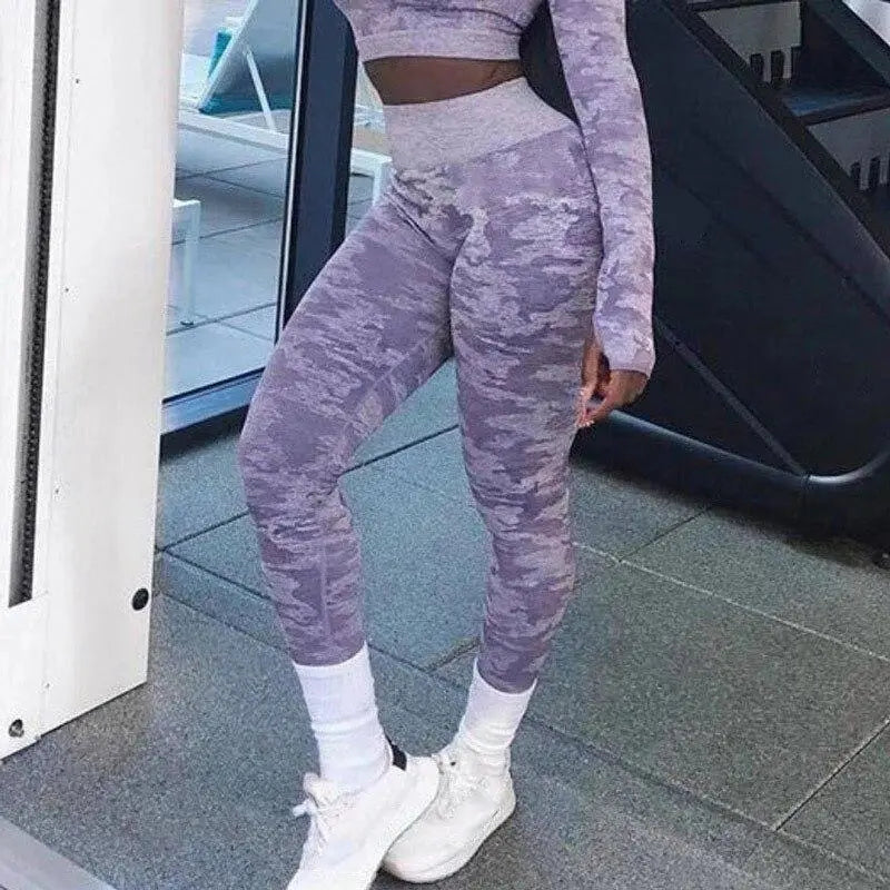 A person wearing purple camo Fitness-8 high waist fitness leggings and white shoes stands near gym equipment.
