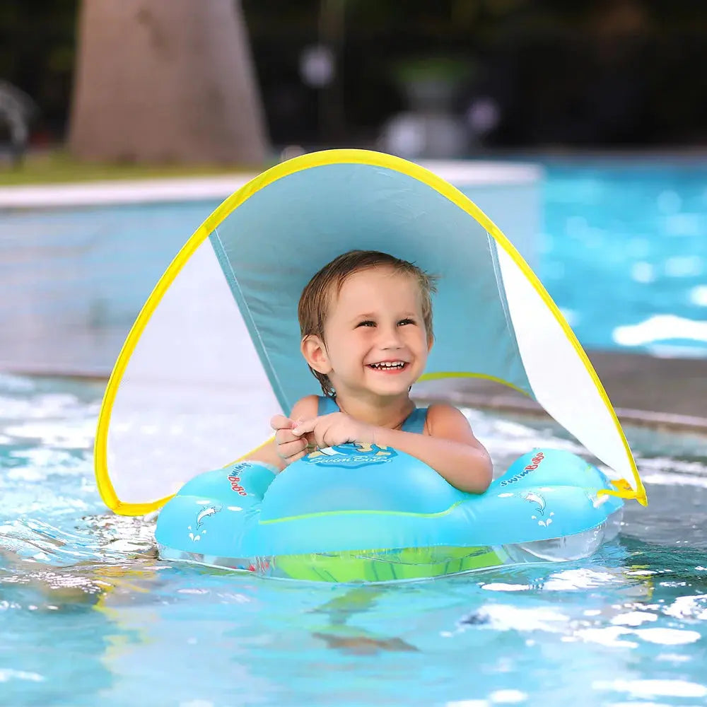 A toddler with a joyful expression floating in a blue and yellow Soulful Trading baby swimming pool float with a detachable sunshade in a swimming pool.