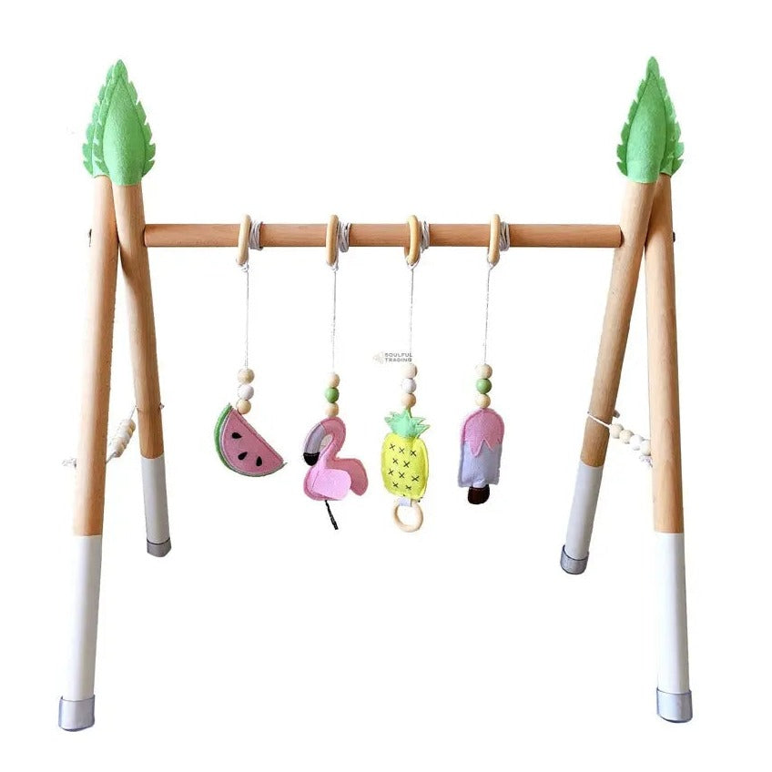 Baby gym with Montessori-inspired design, featuring a wooden frame and hanging toys shaped like watermelon, pineapple, flamingo, and ice cream, adorned with green treetop ornaments - Soulful Trading&