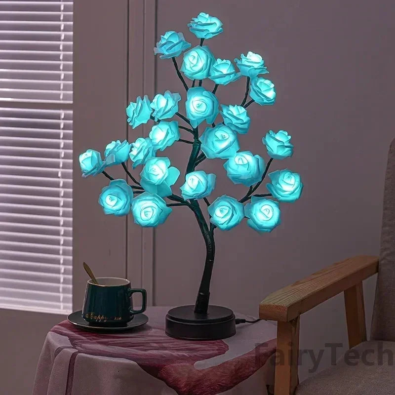 An illuminated Soulful Trading Forever Rose Tree Lamp with blue rose-shaped lights, placed on a side table next to a green mug in a dimly lit room.