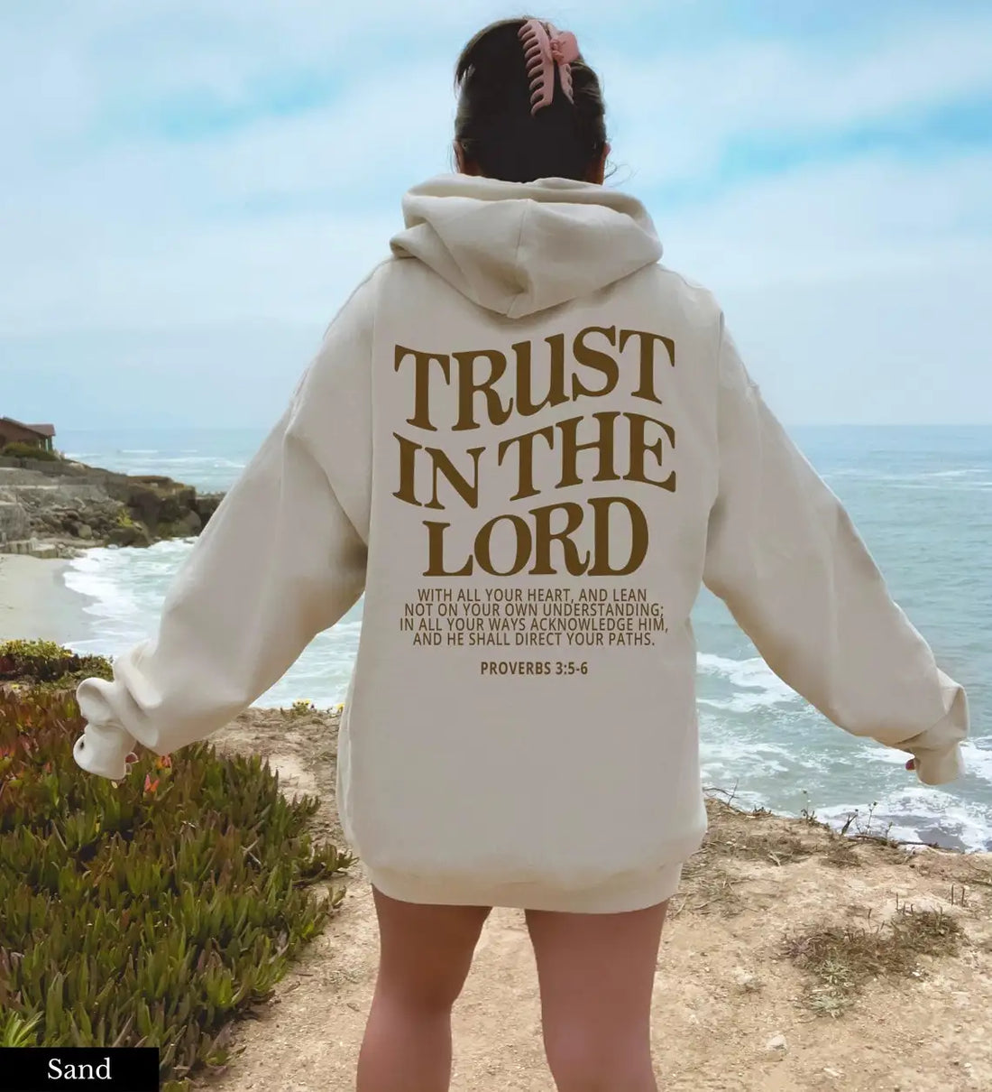 Woman standing on a coastline, wearing a casual hoodie with &quot;Trust In the Lord Christian Sweatshirts&quot; and a Bible verse from Proverbs 3:5-6 printed on the back by Soulful Trading.
