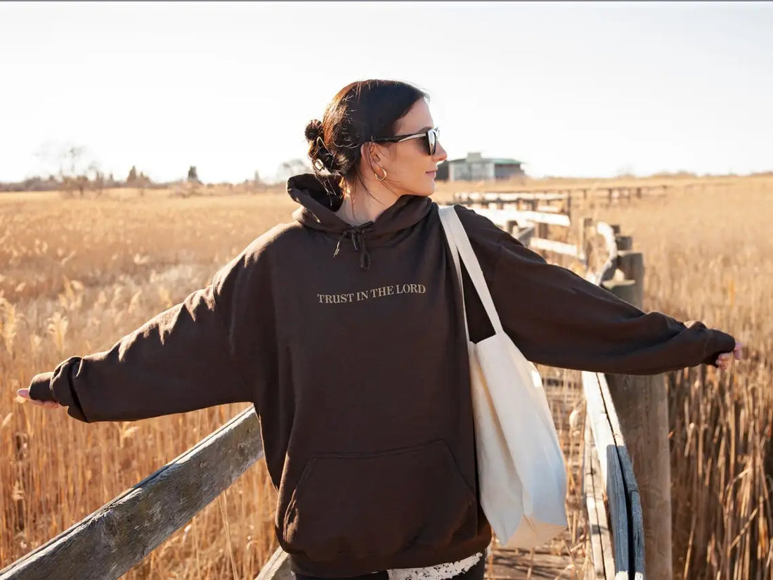 Woman with sunglasses stands on a wooden bridge in a marshy area, wearing a Soulful Trading Trust In the Lord Christian Sweatshirt, arms outstretched, enjoying the sunlight.