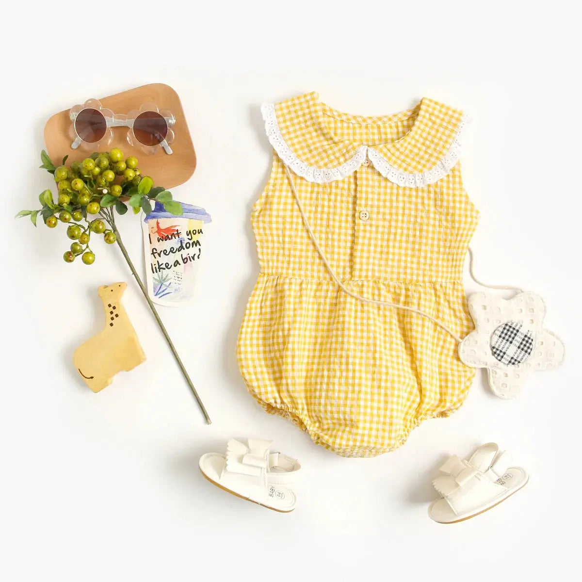 Flat lay of baby items including a Soulful Trading Spring &amp; Summer Baby Onesie, white shoes, sunglasses, a pacifier, and a plush toy on a white background.