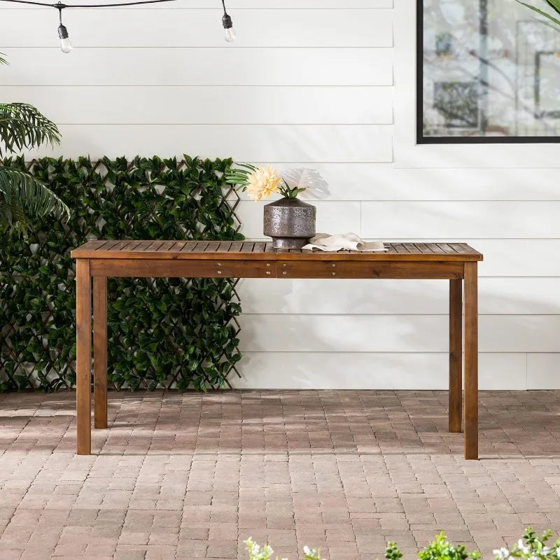 A modern outdoor Crosswinds Patio Dining Table by Walker Edison with a plant in a decorative pot and a sheer cloth, set on a brick patio against a backdrop of a lush green hedge and a white wall.