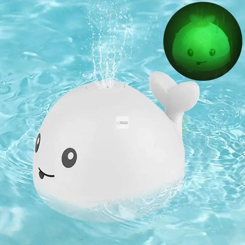A white, Soulful Trading Sprinkler Whale Bath Toy floats in clear blue water, with a small inset showing the toy’s green, glowing light feature.