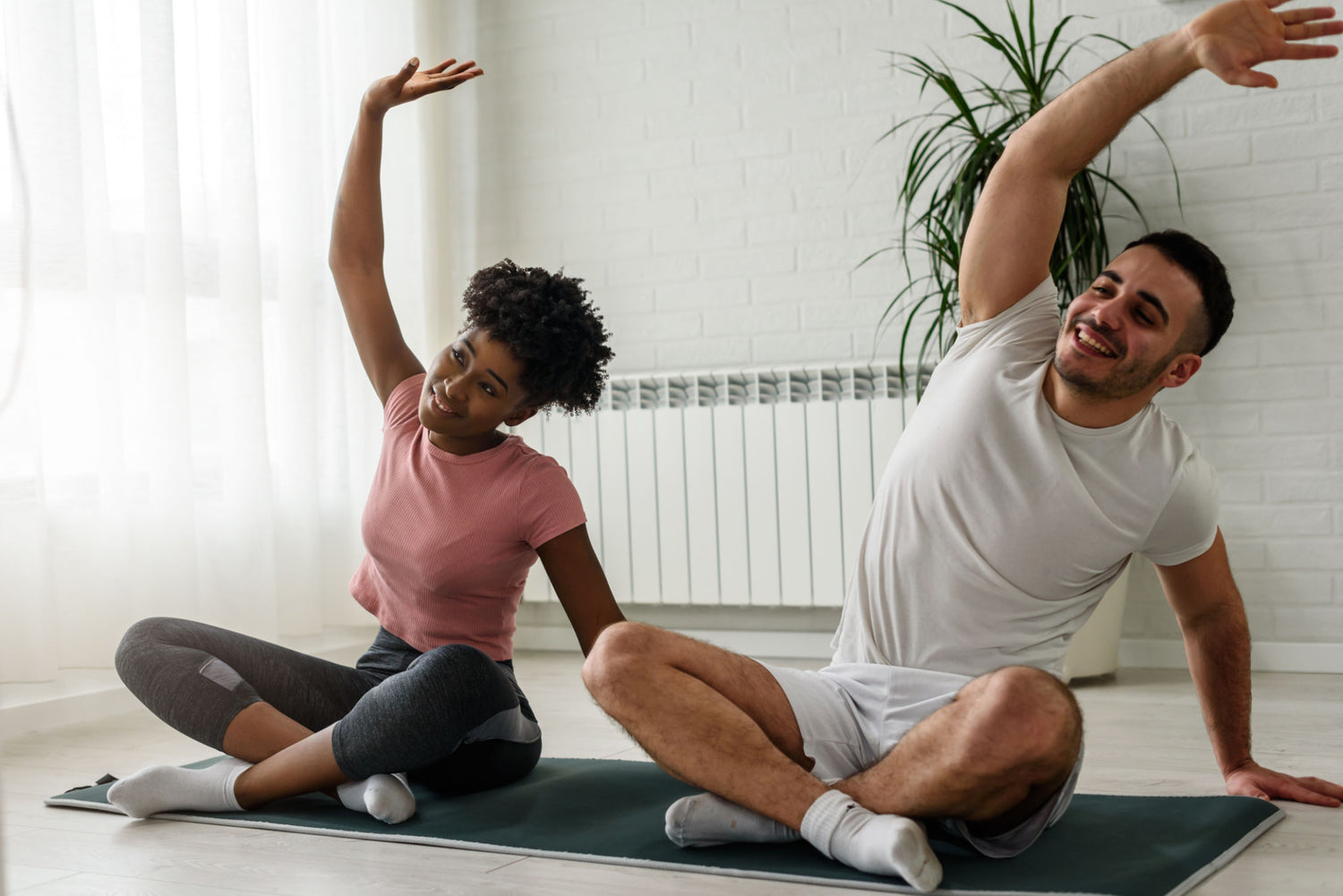A man and a woman sitting on yoga mats, smiling and doing side stretch exercises in a bright room.